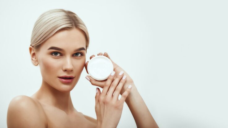 You need a special face cream. Cute young blonde woman showing face cream while standing against